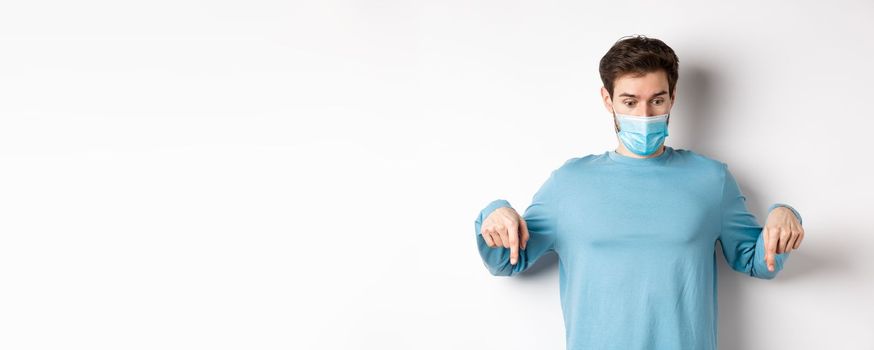 Covid-19, health and quarantine concept. Curious man checking out advertisement, pointing and looking down, wearing medical mask with casual blue sweatshirt, white background.