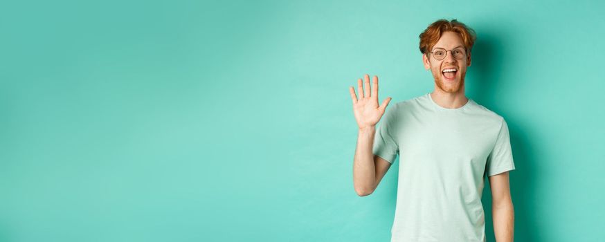 Friendly bearded guy in glasses saying hello, waving hand to greet and welcome you, standing cheerful and smiling over turquoise background.