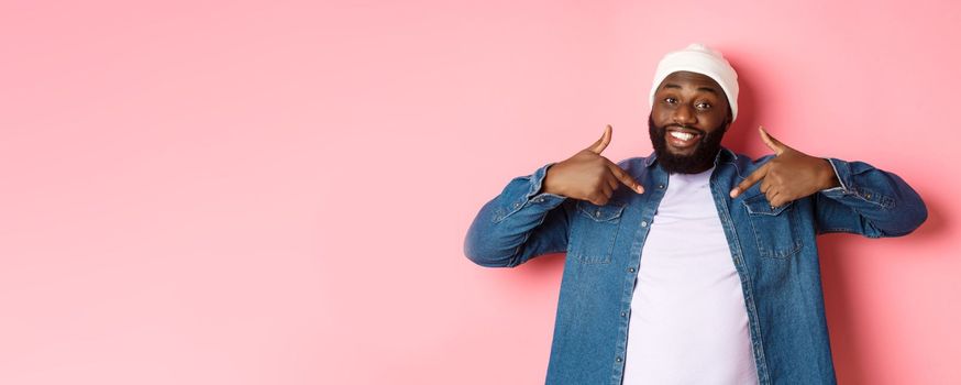 Happy young african-american hipster guy pointing at himself t-shirt, smiling pleased, standing over pink background.