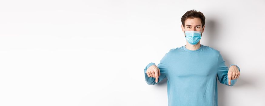 Covid-19, health and quarantine concept. Smiling caucasian man in face mask pointing fingers down, showing logo, standing over white background.