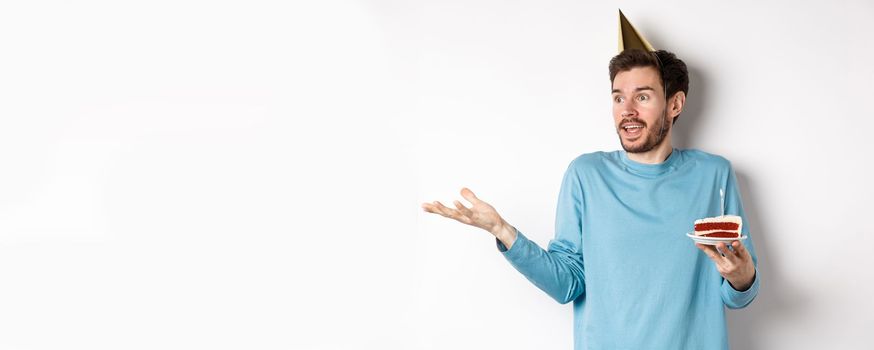 Celebration and holidays concept. Surprised man shrugging in disbelief, looking away, holding birthday cake and wearing party cone hat, white background.