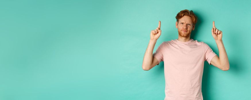 Skeptical and disappointed redhead man pointing fingers up, frowning displeased and looking upset, standing over turquoise background.