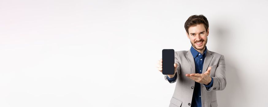 E-commerce and online shopping concept. Here you go. Smiling pleasant salesman demonstrate promo on phone, showing empty smartphone screen, white background.