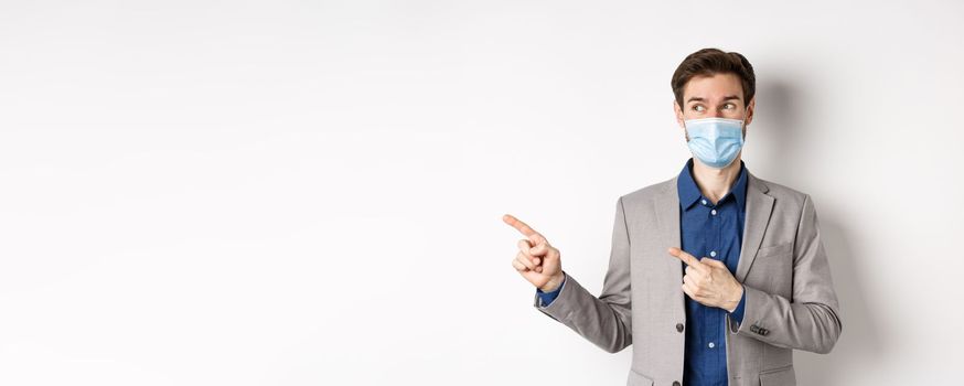 Covid-19, pandemic and business concept. Excited young man in medical mask and suit looking left, pointing at logo with pleased face, white background.