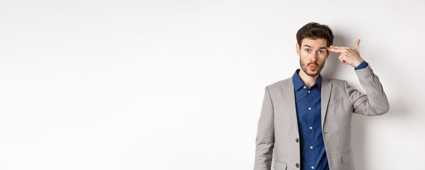 Annoyed guy in business suit shoot himself with hand gun near head, look distressed and tired after work, standing on white background.