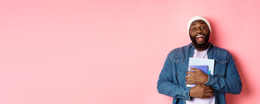 Education. Happy african-american male student in beanie holding notebooks, studying courses, smiling at camera, standing over pink background.