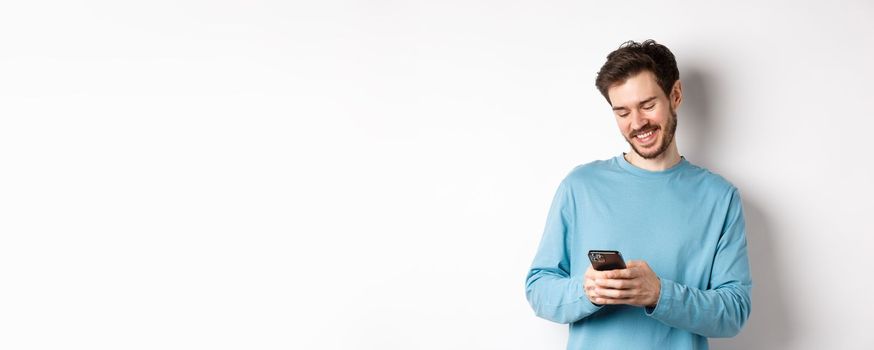 Portrait of modern caucasian man with beard, wearing casual clothes, reading smartphone screen and smiling, networking via mobile phone, standing over white background.