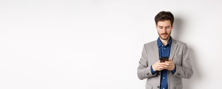 Businessman chatting on mobile phone, standing in suit with serious face, white background.