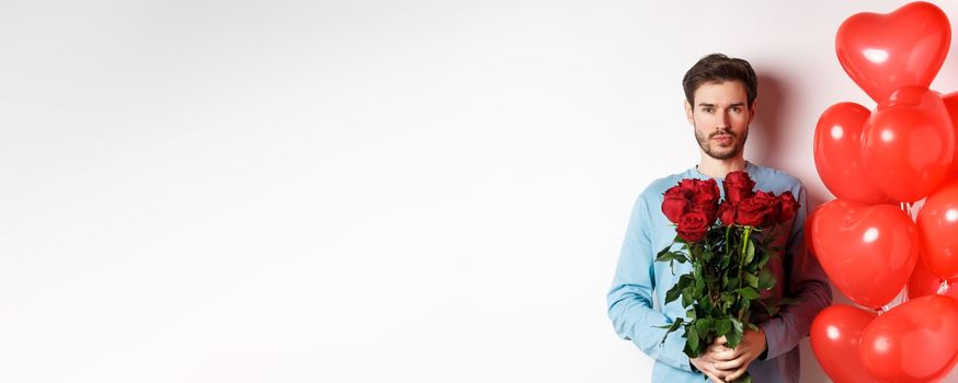 Valentines day romance. Confident young man holding bouquet of red roses, standing near hearts balloons, going on romantic date with lover, white background.