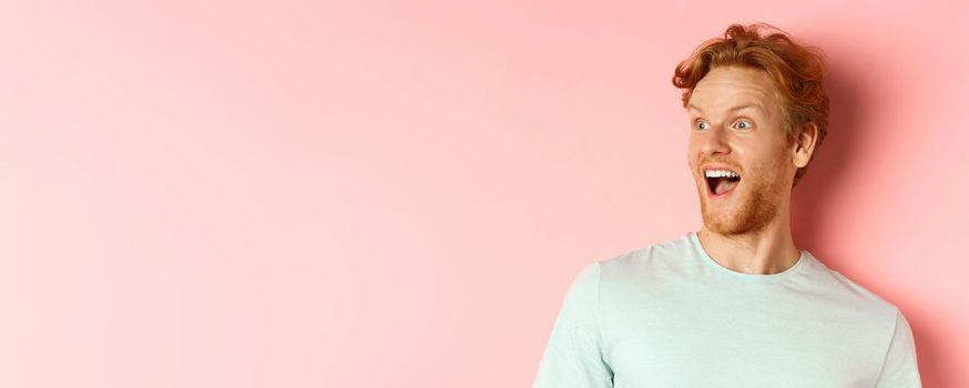 Close up of happy and fascinated young man with red hair checking out promo deal, looking left with amazed smile, standing over pink background.