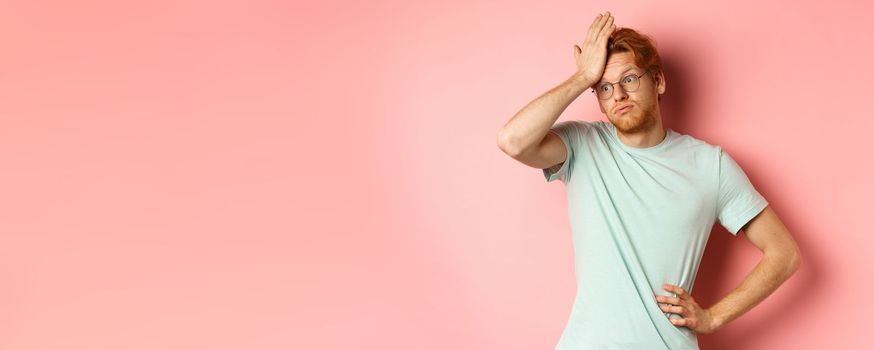 Tired young man with red hair, wearing glasses, looking annoyed and tensed, making facepalm gesture and exhale bothered, standing over pink background.