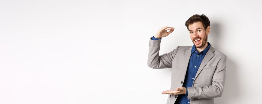 Handsome salesman in suit showing big size, large income, smiling at camera enthusiastic, standing on white background.