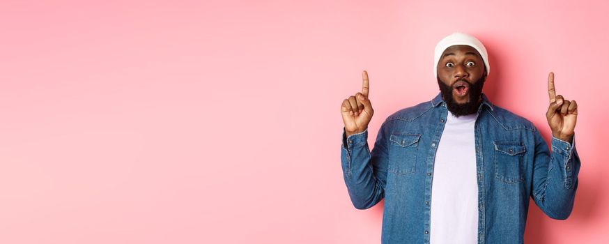 Excited and impressed Black man showing amazing promo, pointing fingers up at logo, standing against pink background.