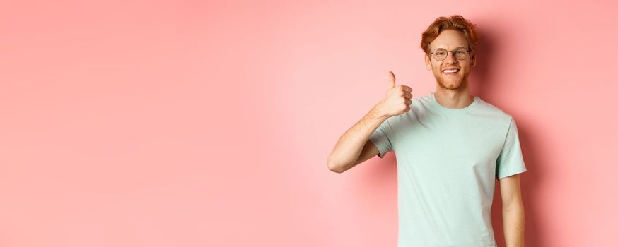 Cheerful redhead man in glasses and t-shirt, showing thumbs up with satisfied face, showing positive reaction, approve and agree with you, standing over pink background.