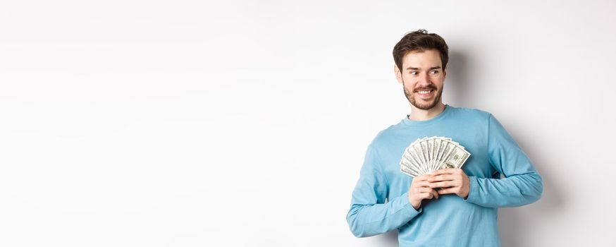 Greedy smiling man showing money and looking right, thinking about shopping, standing over white background.