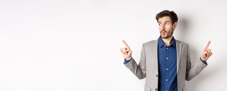 Indecisive male entrepreneur in suit pointing fingers sideways, choosing between two variants, looking at left logo pensive, standing on white background.