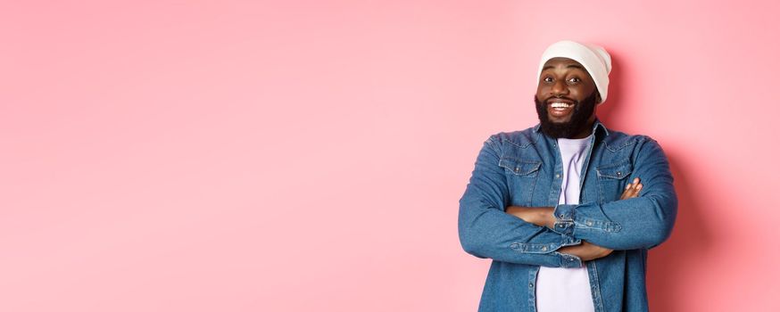 Happy Black man with beard, wearing beanie and denim shirt, looking intrigued and amused at camera, smiling with arms crossed on chest, pink background.