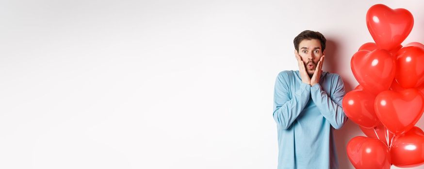 Valentines day. Image of young man standing near hearts balloons with shocked face, staring startled at camera, white background.