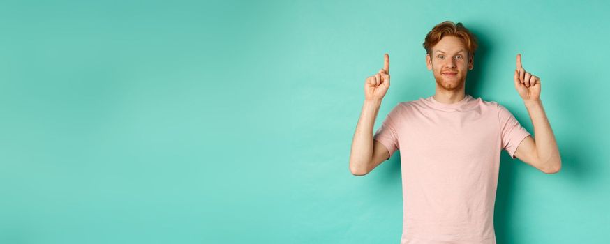 Attractive bearded man with red hair, wearing t-shirt, smiling cheerful and pointing fingers up, showing advertisement, standing over turquoise background.