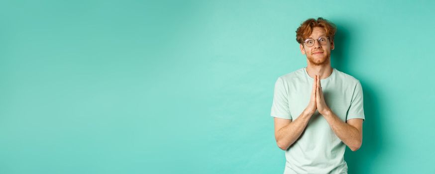 Cute redhead man asking for help, saying please with namaste gesture, pleading or begging for favour, looking silly at camera, standing over mint background.