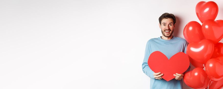 Smiling boyfriend holding Valentines card and standing near romantic red heart balloons, celebrating lovers day, standing over white background.