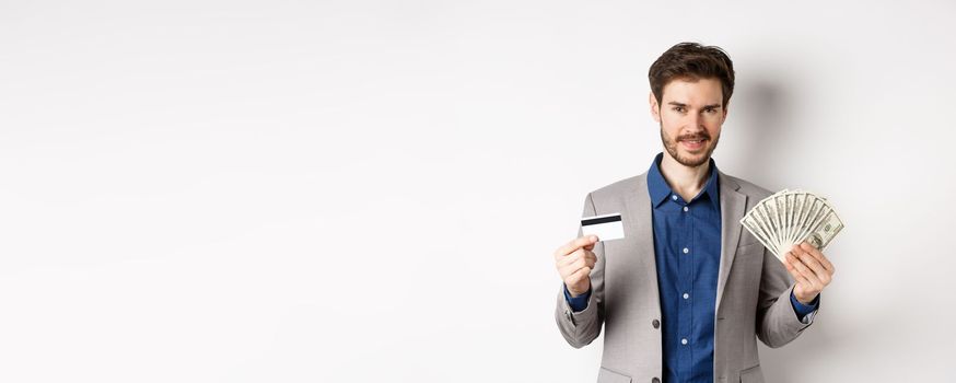 Rich smiling man in suit showing money and plastic credit card, standing with dollar bills and look pleased, white background.