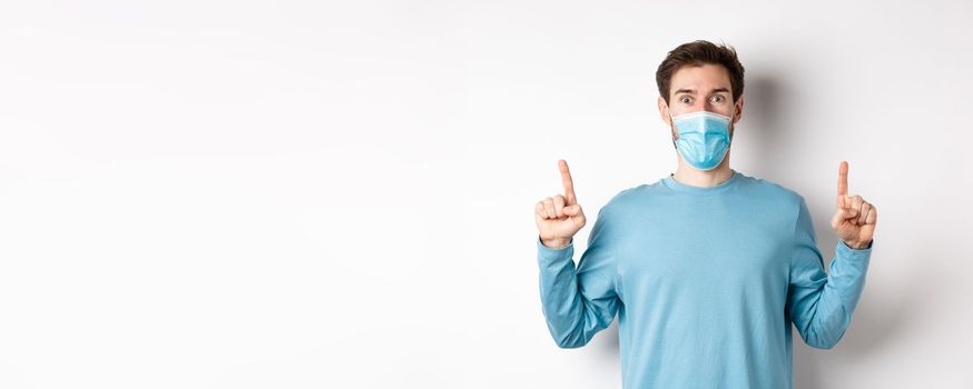 Coronavirus, health and quarantine concept. Excited smiling guy checking out special deal, pointing fingers up, stare amazed at camera, wear medical mask on white background.