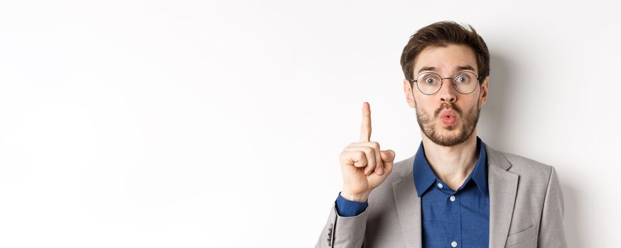 Excited businessman in glasses and suit pitching idea, raising finger and say suggestion, have a plan, standing against white background.