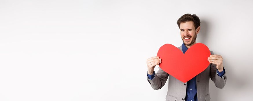 Charismatic young man winking and smiling, showing big red heart cutout for Valentines day date, say love you to lover, standing over white background.