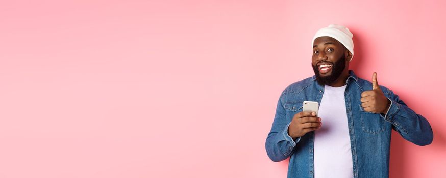 Technology and online shopping concept. Satisfied Black man smiling, using mobile phone and showing thumb-up in approval, making order, standing over pink background.