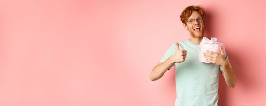 Valentines day and romance concept. Cheerful young man holding box with gift and showing thumbs-up, thanking for present, standing over pink background.