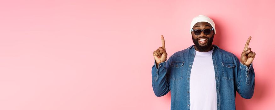 Happy bearded african-american man showing announcement, pointing fingers up and smiling, standing in beanie and sunglasses over pink background.