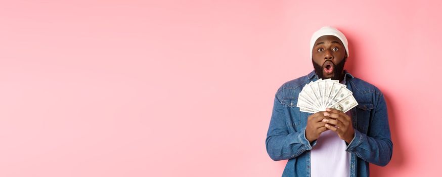 Amazed african-american man receive cash prize, showing money and staring at camera in awe, standing over pink background.