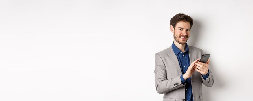 Smiling successful business man using smartphone on white background, standing in suit.