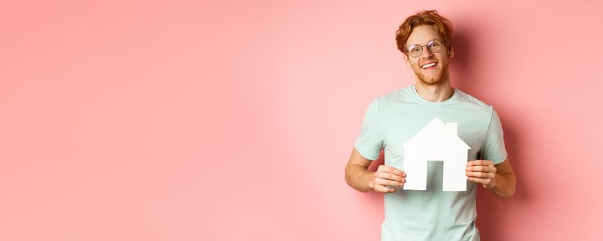 Real estate. Cheerful young man with red hair, wearing glasses and t-shirt, showing paper house cutout and smiling, buying apartment, pink background.