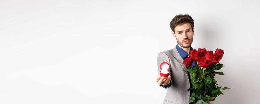 Handsome boyfriend in suit asking to marry him, standing with red bouquet of roses and engagement ring, looking pleading at camera, standing over white background.