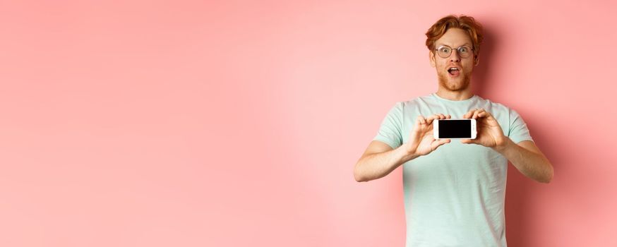 Amazed redhead man gasping and staring with awe at camera, showing blank smartphone screen horizontally, standing over pink background.