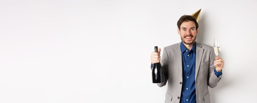 Celebration and holidays concept. Excited handsome birthday guy in party hat smiling, drinking champagne and having fun, white background.