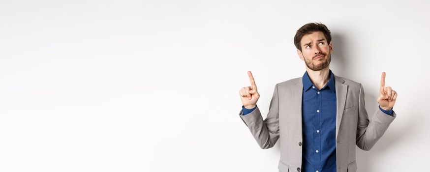 Skeptical and hesitant young businessman in suit looking up with disappointed smirk, feel unsure about deal, standing dissatisfied on white background.