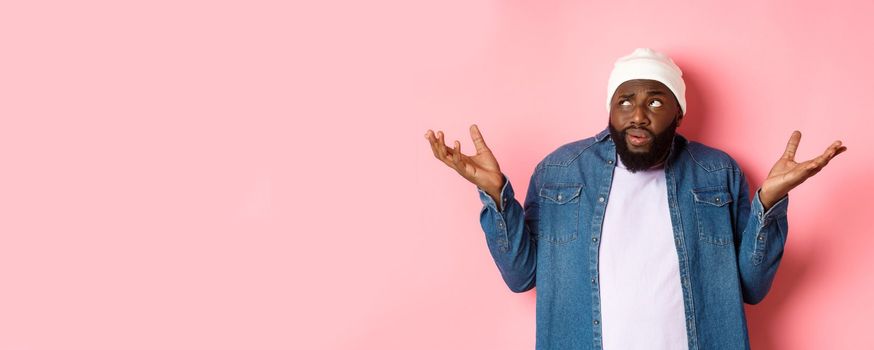 Confused african-american man in beanie looking at upper left corner doubtful, shrugging uncertain, standing over pink background.