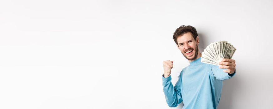 Happy caucasian man stretch out hand with money in dollars, saying yes and celebrating income, got cash prize, standing over white background.