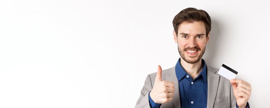 Shopping. Handsome satisfied client showing thumbs up and plastic credit card, smiling pleased, white background.