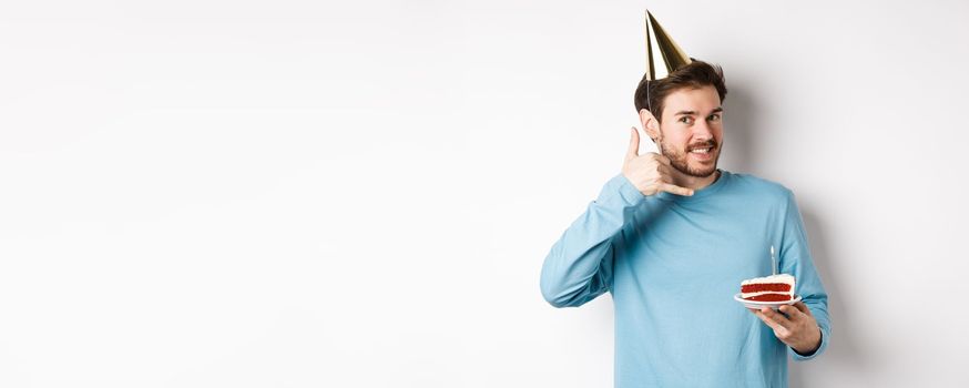 Celebration and holidays concept. Cheerful smiling man in party hat, celebrating birthday with bday cake, showing call me phone gesture near ear, white background.