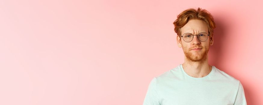 Headshot of skeptical redhead man in glasses and t-shirt frowning disappointed, staring with disapproval and judgement, standing over pink background.