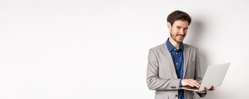 Successful smiling businessman working on laptop and looking happy at camera, standing in grey suit on white background.