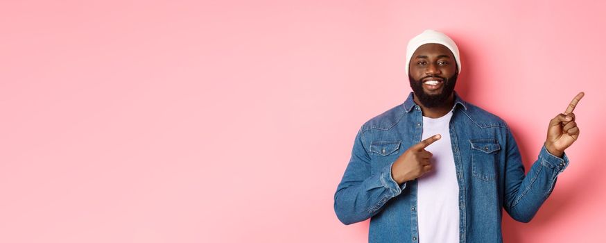 Happy african-american man smiling, pointing fingers right and showing promo, making announcement, standing over pink background.