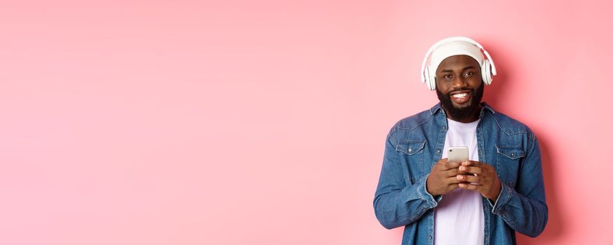 Image of handsome Black guy in headphones, listening music and using mobile phone, smiling at camera, pink background.