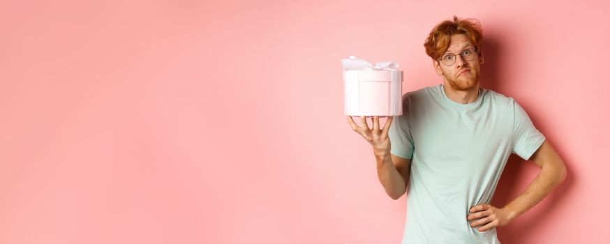 Love and holidays concept. Funny redhead guy shrugging silly and showing pink gift box for valentines day, standing over pink background.