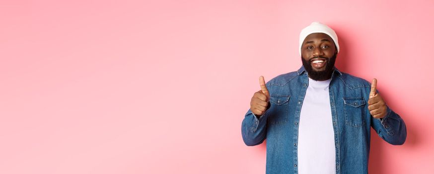 Happy african-american man showing thumbs-up in approval, like something good, praising great deal, standing over pink background.