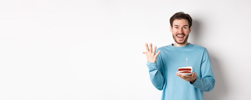 Excited man receive birthday surprise, holding bday cake and smiling happy, standing over white background, making wish on lit candle.